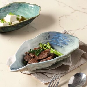 Può includere: Una ciotola in ceramica irregolare a forma di conchiglia con una finitura smaltata reattiva lucida blu chiaro e bianca. La ciotola contiene pezzi di bistecca cotta e asparagi verdi, poggiata su un tovagliolo di lino beige con forchetta e cucchiaio.