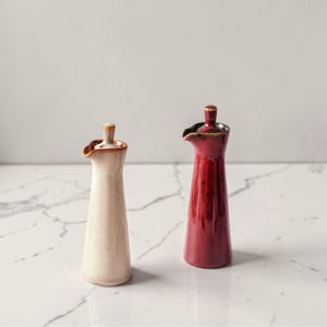 Puede incluir: Dos dispensadores de aceite de cerámica. Uno es de color blanco crema y el otro rojo intenso. Ambos tienen una forma delgada y alargada con un pico y un pequeño tapón redondeado. Los dispensadores son probablemente para uso en la cocina.
