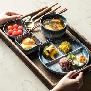 Puede incluir: Una bandeja de madera presenta una comida completa con varios platos. La comida incluye un cuenco de sopa, un plato dividido con verduras y cereales, y pequeños cuencos de tomates y otros acompañamientos. También se incluyen palillos y cucharas.