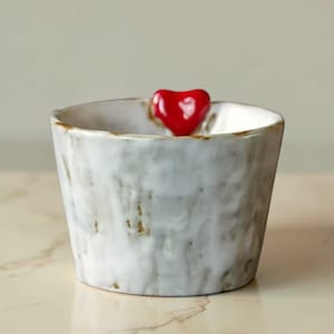 Può includere: Una piccola ciotola in ceramica bianca con una superficie strutturata e un accento a forma di cuore rosso. La ciotola ha un aspetto leggermente ruvido, fatto a mano, con sottili accenti marroni sul bordo.
