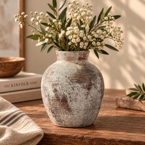May include: A round ceramic vase with a distressed, weathered finish in shades of white and brown. The vase has a wide body and a narrow neck, filled with a bouquet of small white flowers and green leaves, creating a textured, rustic appearance.