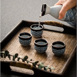 May include: A sake set with a ceramic bottle pouring liquid into three small cups. The cups and bottle have a blue wave pattern and brown trim. The set is on a woven tray.