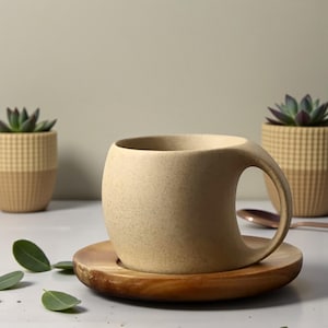 Può includere: Una tazza in ceramica beige con un'esclusiva maniglia curva è appoggiata su un piattino di legno. La tazza ha una consistenza macchiettata e un'ampia apertura. Un cucchiaio di rame e due piccole piante grasse in vaso sono sullo sfondo.