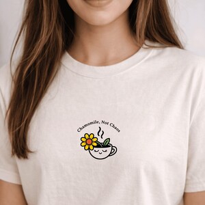 以下が含まれることがあります： 「Chamomile, Not Chaos」の文字と、カモミールのお花のイラストが描かれたクリーム色のTシャツ。黄色い花びらとオレンジ色の中心を持つカモミールのお花と、目を閉じているティーカップが特徴です。