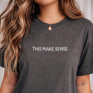 May include: Dark gray t-shirt with the white text "THIS MAKE SENSE" centered on the front. The shirt has short sleeves and a crew neck. The model is wearing a gold necklace.