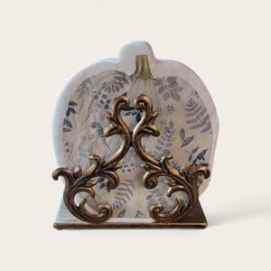 May include: A decorative napkin holder with a pumpkin-shaped back. The holder features a gold-colored, ornate scrollwork design in the front. The napkins have a blue and white floral pattern.
