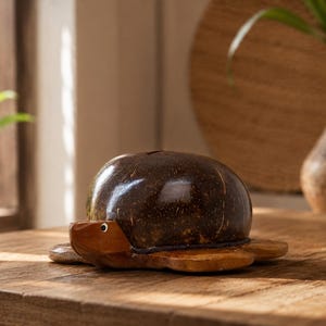 May include: A handcrafted turtle-shaped piggy bank made from a coconut shell and wood. The dark brown shell forms the turtle's body, while the wooden base and head are a lighter brown. The piggy bank sits on a wooden table, with a decorative vase in the background.