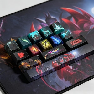 Könnte beinhalten: Individuelle Tastenkappen mit League of Legends-Grafiken. Die Tastenkappen zeigen verschiedene Charakterdesigns und Aktionssymbole in Blau-, Grün-, Rot- und Gelbtönen. Die Leertaste zeigt eine große Grafik eines Charakters. Auf einer Tastenkappe steht "LEAGUE OF LEGENDS".