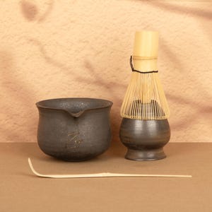 Op de afbeelding: Een matcha-theeset met een donkergrijze keramische kom met een tuit, een bamboe klopper en een bamboe lepel. De set is geplaatst op een bruin oppervlak met een neutrale achtergrond.