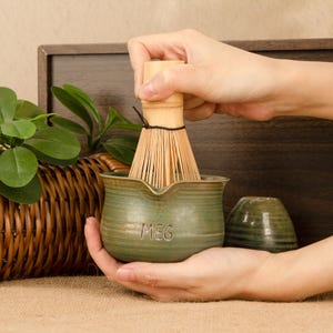 May include: A matcha tea set featuring a green ceramic bowl and a bamboo whisk. The bowl is engraved with the name "MEG". The whisk is in use inside the bowl. A small, matching green ceramic cup is also visible.