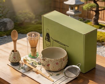 May include: A matcha tea set including a light green box with black line art, a ceramic bowl with a floral pattern and the word Tea, a bamboo whisk in a clear case, a bamboo scoop, a ceramic scoop holder, and a metal mesh strainer on a cloth mat.