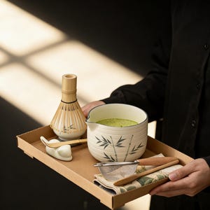 May include: A matcha tea set on a cardboard tray. The set includes a bamboo whisk, a ceramic bowl with a bamboo design, a strainer, a spoon, and a small ceramic whisk holder. The bowl is filled with green matcha tea.