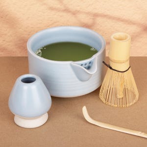 May include: A light blue ceramic matcha bowl with a spout, filled with green matcha tea. A bamboo whisk, a matcha spoon, and a light blue ceramic matcha bowl rest are also present. The items are arranged on a brown surface.