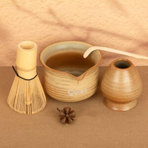 May include: A matcha tea set comprising a bamboo whisk, a ceramic bowl with the word "Quiet", a ceramic tea bowl, and a wooden scoop. The set is in beige and brown tones, with a small pine cone.
