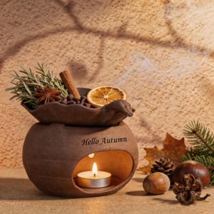 Peut inclure: Un brûle-parfum en céramique brune avec l'inscription Hello Autumn gravée sur le devant. Le bol supérieur contient des brins de romarin, un bâton de cannelle, de l'anis étoilé, des clous de girofle et une tranche d'orange séchée.