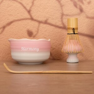 May include: A ceramic matcha bowl with a pink and white ombre finish and the word Harmony printed in white, accompanied by a light brown bamboo matcha whisk and a long, curved bamboo tea scoop on a flat surface.