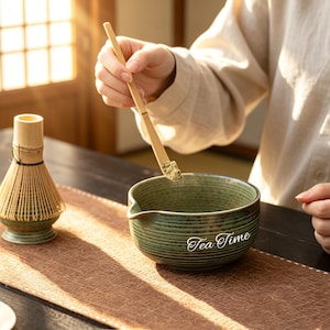 May include: A green ceramic tea bowl with the text "Tea Time" is being prepared with matcha powder. A bamboo whisk and scoop are also present, suggesting a tea ceremony. The bowl is approximately 10 cm in diameter.