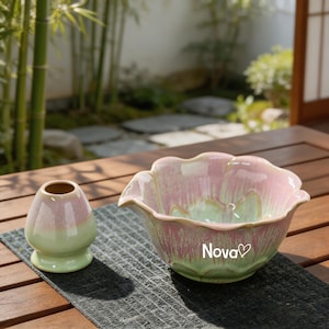 May include: A ceramic matcha bowl and a matching whisk holder featuring a scalloped rim and a gradient glaze of soft pink and pale green. The bowl displays the text Nova with a heart symbol printed in white on the exterior surface.