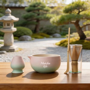 May include: A matcha tea set featuring a pink and green gradient ceramic bowl with the text Matcha life, a matching pink and green whisk holder, a bamboo scoop, and a bamboo whisk inside a clear cylindrical container on a wooden surface.