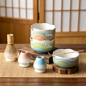 May include: A selection of ceramic matcha tea bowls and accessories. The bowls are painted with pastel colours and the words "Zen" and "Calm". Includes a bamboo whisk, two small holders, and a wooden scoop.