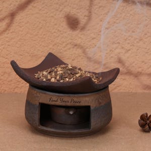 Puede incluir: Un quemador de aceite de cerámica marrón oscuro con un plato superior ancho y curvado lleno de trozos botánicos secos. La base presenta una abertura para una vela de té y el texto Find Your Peace impreso en la parte frontal.