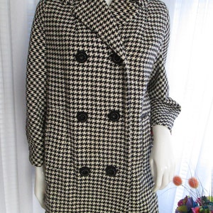 May include: A black and white houndstooth patterned coat with a double breasted closure and black buttons. The coat has a collar and long sleeves.