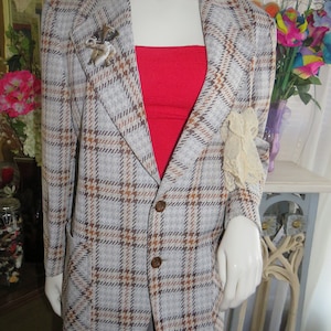 Può includere: Un blazer a quadri marrone, bianco e nero con una camicetta rossa sotto. Il blazer ha una tasca sul lato sinistro e due bottoni sul davanti. C'è una spilla sul risvolto sinistro.