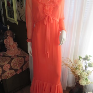 May include: A long, flowing, coral-colored dress with a ruffled neckline and a tie at the waist. The dress has long sleeves and a flowing skirt with a ruffled hem.