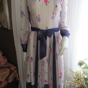 Puede incluir: Un vestido blanco con una cinta azul marino y una falda plisada. El vestido tiene un fondo blanco con un patrón de lunares rosas, morados y azules.