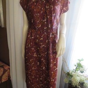 Op de afbeelding: Een vintage jurk uit de jaren 1950 met een marroon en oranje bloemenprint. De jurk heeft een ronde hals, korte mouwen en een getailleerd bovenlijf. Het heeft een decoratieve broche op de halslijn.