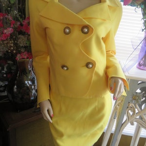 May include: A yellow skirt suit with a double-breasted jacket and gold buttons. The jacket has a wide collar and the skirt is knee-length.
