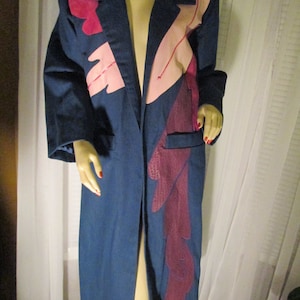 May include: A long, blue denim coat with pink and purple leather applique details. The coat has a long, open front and a single pocket on the left side.