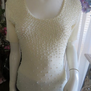 以下が含まれることがあります： 丸首と半袖の白いニットトップス。トップスは小さな白い真珠のようなビーズで覆われています。