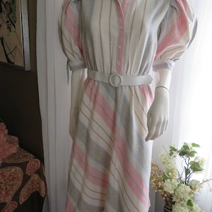 Könnte beinhalten: Ein gestreiftes Kleid in Rosa, Weiß und Blau mit einem weißen Gürtel. Das Kleid hat einen Kragen und kurze, bauschige Ärmel.