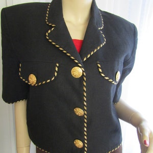 May include: A black cropped jacket with gold trim and large gold buttons. The jacket has short sleeves and a collar.