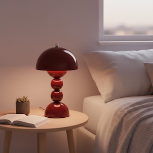 May include: A red table lamp with a dome-shaped shade and a stacked sphere base. The lamp sits on a round wooden side table next to a bed. A small succulent in a pot and an open book are also on the table.