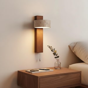 May include: A modern wall sconce with a rectangular wooden base and a cylindrical lampshade. The lampshade is a light beige color with a speckled texture. A pull chain hangs below the wooden base. The sconce is mounted on a light-colored wall.