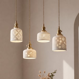 May include: A set of four hanging pendant lights featuring white ceramic shades with various intricate cut-out patterns. Each shade is suspended by a twisted brown fabric cord and attached to a brass-toned metal fixture at the top.