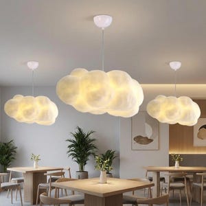May include: Three cloud-shaped pendant lights illuminate a room. The lights are white and fluffy, resembling clouds, and hang from the ceiling. The room features wooden tables, chairs, and potted plants, creating a cozy atmosphere.