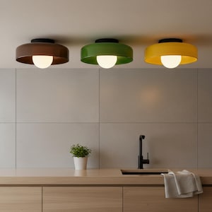 May include: Three modern ceiling lights in a row, each with a different colored shade: brown, green, and yellow. Each light has a black base and a white, spherical bulb. The lights are mounted on a white ceiling.