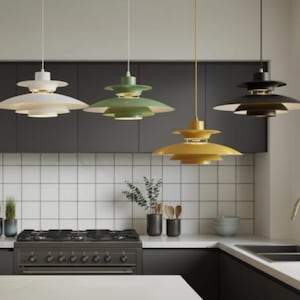 May include: Four modern pendant lights in a kitchen setting. The lights are multi-tiered, with a white, green, yellow, and black finish. The kitchen features a stove, white tile backsplash, and dark cabinets.