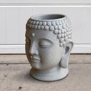 May include: A gray Buddha head planter with closed eyes and detailed hair. The planter has a wide opening at the top and a rounded base. The surface has a speckled texture, and the overall design is serene and contemplative.