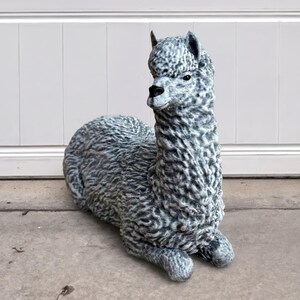 May include: A gray and white alpaca statue in a resting position. The alpaca has a textured, woolly appearance and is detailed with a black nose and eyes. The statue is made of a durable material and is suitable for outdoor display.