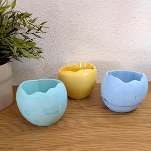 Peut inclure: Trois petits pots de fleurs ronds en forme d'œuf avec des bords festonnés et irréguliers. Un pot est bleu clair avec un marbrage blanc, un est jaune uni et un est bleu plus foncé. Chaque récipient a une finition mate lisse et un centre creux.