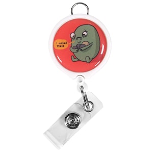 May include: A white and red retractable badge reel with a cartoon illustration of a green character holding purple objects. The character has a speech bubble that says "I maked these". The reel has a metal clip and a clear plastic strap.