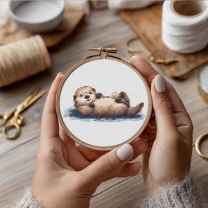 Peut inclure: Un cercle de broderie au point de croix représentant une loutre brune allongée sur le dos. La loutre est brodée dans des tons de marron et de beige, avec un fond bleu. Le cercle est tenu par des mains, et le cadre en bois est visible.