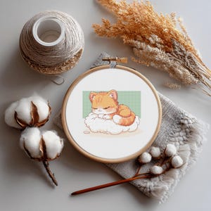 Puede incluir: Un bastidor de bordado de punto de cruz con un gato atigrado naranja durmiendo sobre una nube. El bordado está en un bastidor de madera, sobre un paño gris. Otros elementos incluyen capullos de algodón, hilo y trigo seco.