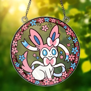 以下が含まれることがあります： ピンク、白、青を基調とした、漫画のキャラクター、Sylveonが描かれた丸いステンドグラスの窓。 デザインはピンクと青の花の縁で囲まれ、銀色のチェーンで吊り下げられています。