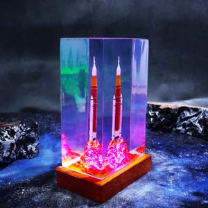 May include: A clear resin sculpture featuring two detailed rocket models with orange and white accents, set against a vibrant, colorful backdrop. The rockets appear to be launching, with a fiery, illuminated base, mounted on a wooden base.