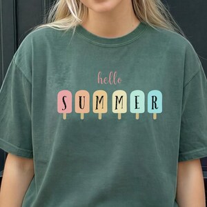 May include: Dark green t-shirt with the words "hello SUMMER" printed on the front. The word "hello" is in a cursive font, and "SUMMER" is in a block font with colorful popsicle graphics. The shirt is a casual, oversized style.